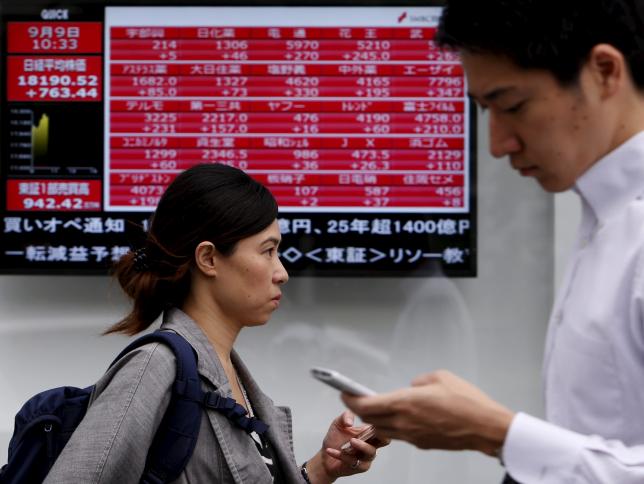 Pedestrians holding their mobile phone walk past an electronic board showing the various stock prices outside a brokerage in Tokyo, Japan, September 9, 2015. REUTERS/Yuya Shino