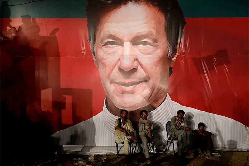 FILE PHOTO: Labourers, who set up the venue, sit under a wall with a billboard displaying a photo of Imran Khan, chairman of the Pakistan Tehreek-e-Insaf (PTI), political party, as they listen to him during a campaign rally ahead of general elections in Karachi, Pakistan July 22, 2018. Photo: Reuters