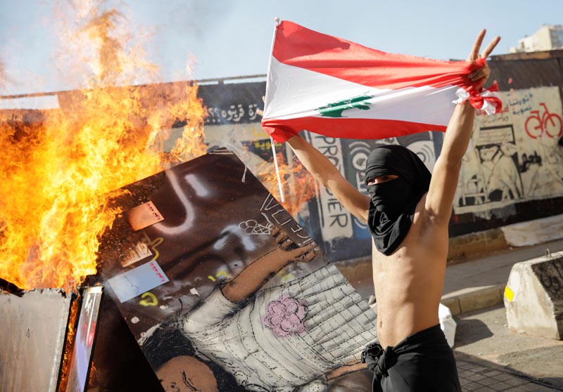 A masked protester holding a Lebanese flag walks past a burning barricade during ongoing anti-government protests in Beirut, Lebanon November 19, 2019. Photo: Reuters