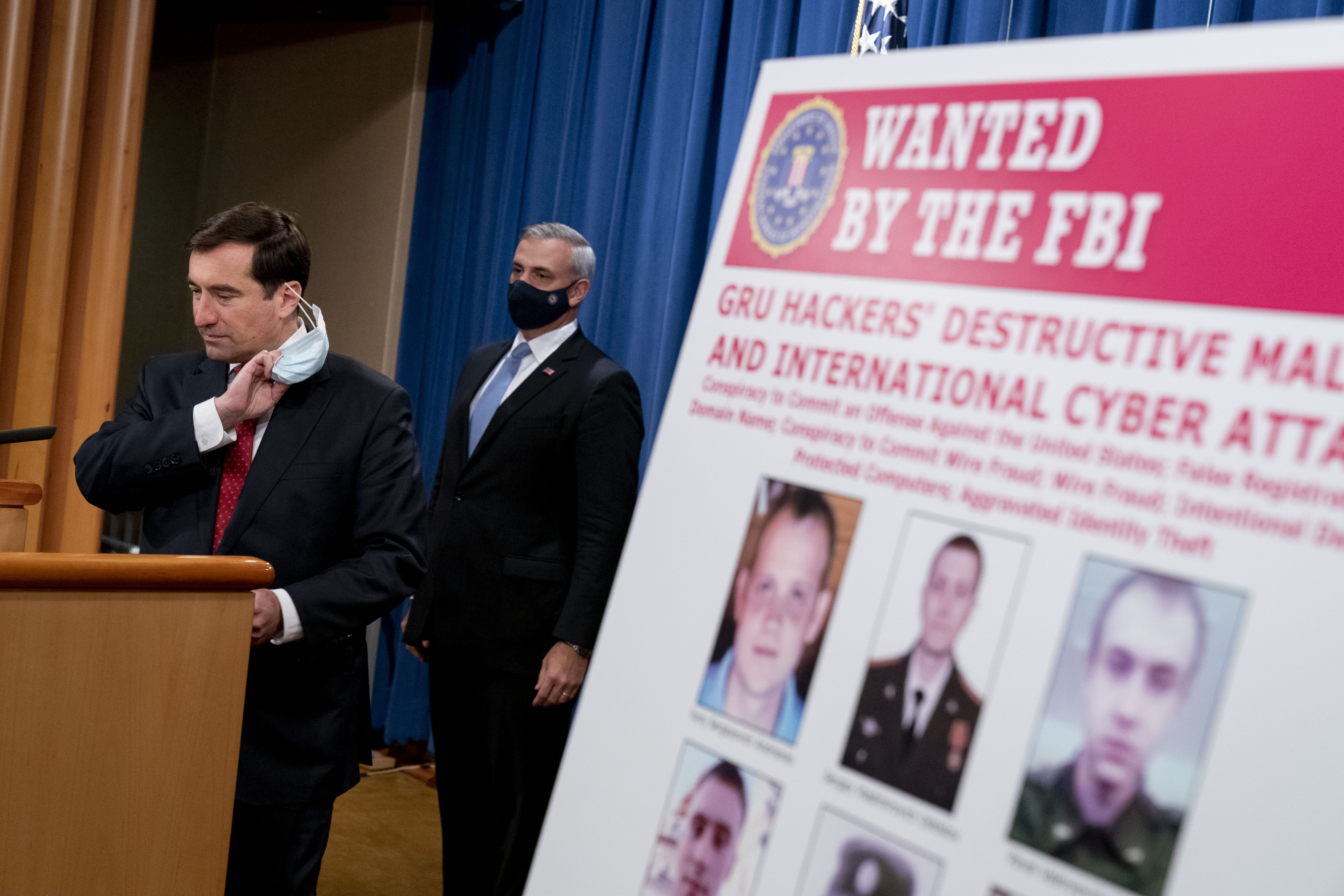 A poster showing six wanted Russian military intelligence officers is displayed as Assistant Attorney General for the National Security Division John Demers, left, takes the podium to speak at a news conference at the Department of Justice, Monday, Oct. 19, 2020, in Washington. Also pictured is US Attorney for the Western District of Pennsylvania Scott Brady, center. (AP Photo/Andrew Harnik, pool)